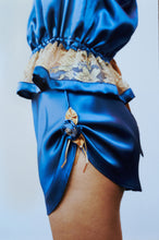 Load image into Gallery viewer, side view of a woman wearing the Anna tap pants with the Anna camisole. the sides of the tap pants are swagged up on a thin silk loop decorated at the edge wth a medium sized blue rose with gold ribbon leaves and tipped with pearls.