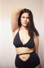 Load image into Gallery viewer, A woman posing with her right hand on the back of her head. She wears the Artemis triangle shaped bralette in petrol blue bamboo jersey. Also the Artemis high waist knickers which have a triangle cut out at the belly button. The jersey on both is trimmed with black elastics and all the hardwear is gold.