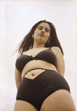 Load image into Gallery viewer, A woman outside against a cloudy sky looking down at the camera. She is wearing the Artemis bralette and high waist knickers. Both are in petrol blue bamboo jersey with black elastics and gold hardware. The front of the knickers has a cross over panel detail that creates an upside down triangle at the waistline around the belly button. The waistband is adjustable above the triangle and has a gold slider.