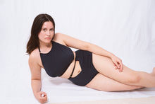 Load image into Gallery viewer, A woman lies on her side looking at the camera. She has long brown hair. She wears the Artemis high neck halter bralette and high waist knickers. The knickers have an upsidedown triangle cut out at the waistline around the belly button. The bralette sits just below her collar bones with a scooped neck.