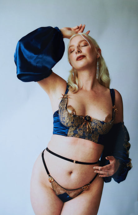 A woman viewed from the front wearing the Anna bra, thong and sleeves, she holds her hand over her head. The blue silk of the sleeve drapes down.