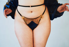 Load image into Gallery viewer, A front vie of the Anna thong. The front has black strapping, gold floral lace, and the blue silk is shaped in a triangle at the front. The sides are decorated with gold ribbon leaves and pearls.
