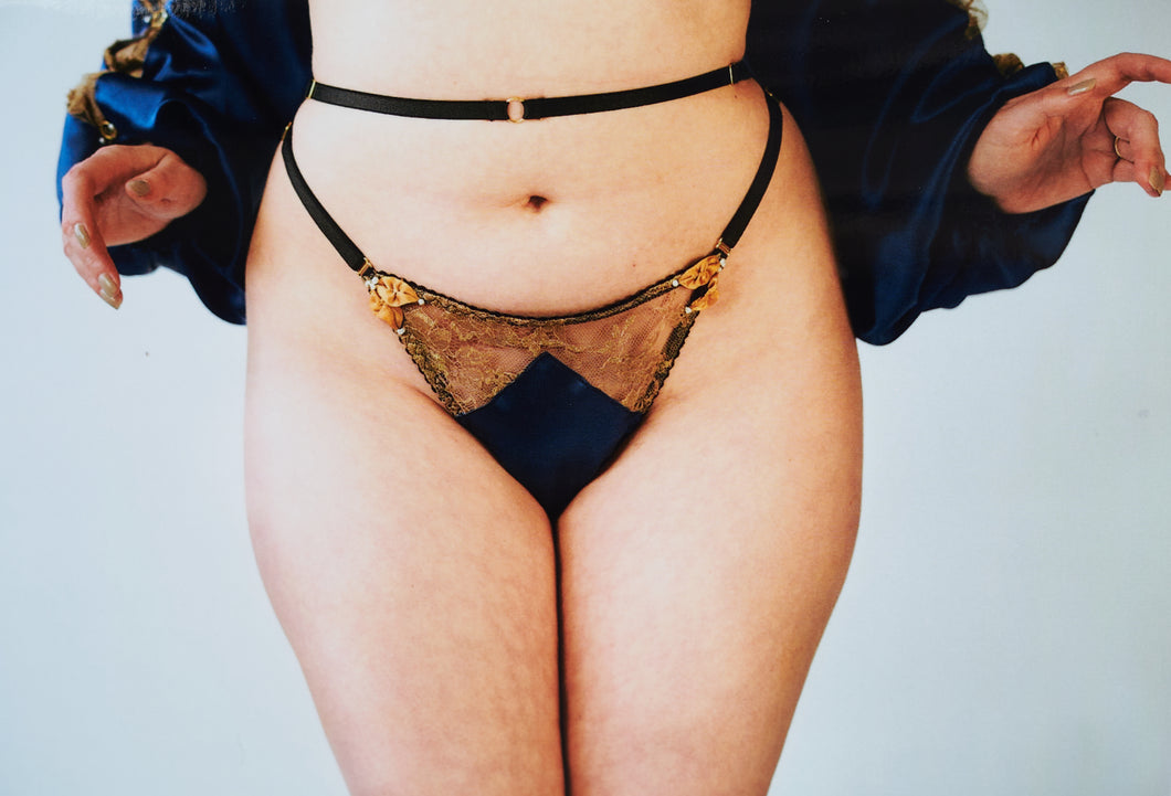 A front vie of the Anna thong. The front has black strapping, gold floral lace, and the blue silk is shaped in a triangle at the front. The sides are decorated with gold ribbon leaves and pearls.