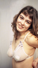 Cargar imagen en el visor de la galería, A model smiles at the camera, the side of the Lily bralette is visible, showing the gold ring that attaches the band strap to the cup. The elastics are off white, there is no frill on the side of the cup.
