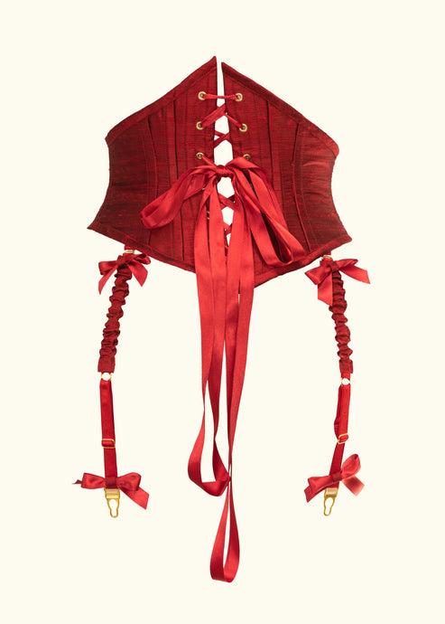 The red silk douppion corset belt from the back. The lacing is red ribbon and there are 2 visible suspenders hanging down from the bottom. The clips are gold and are trimmed at the top and bottom of the straps with red ribbon bows.