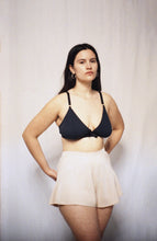 Cargar imagen en el visor de la galería, A woman with long brown hair stands looking at the camera with her hands behind her back. She wears the Artemis bamboo bralette and cacao bamboo tap pants. They flare out from the waistband over her hips to the top of her thighs.
