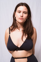 Cargar imagen en el visor de la galería, A close up showing a woman from the waist up looking into the camera. She has long brown hair, gold hooped earrings and wears the Artemis triangle shaped bralette in petrol blue bamboo jersey. The jersey is trimmed with black elastics and a black bow at the front.
