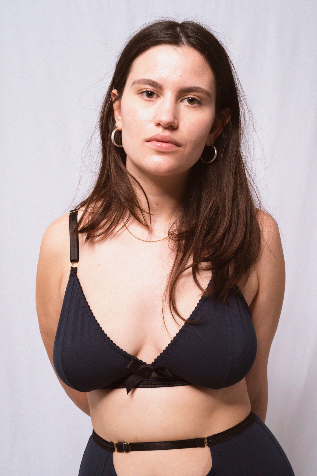 A close up showing a woman from the waist up looking into the camera. She has long brown hair, gold hooped earrings and wears the Artemis triangle shaped bralette in petrol blue bamboo jersey. The jersey is trimmed with black elastics and a black bow at the front.