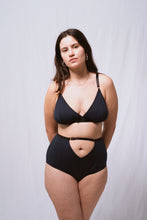 Cargar imagen en el visor de la galería, A long shot of a woman looking at the camera, wearing the Artemis bralette and high waist knickers. Both are in petrol blue bamboo jersey with black elastics and gold hardware. The front of the knickers has a cross over panel detail that creates an upside down triangle at the waistline around the belly button. The waistband carries on across the top of the triangle.
