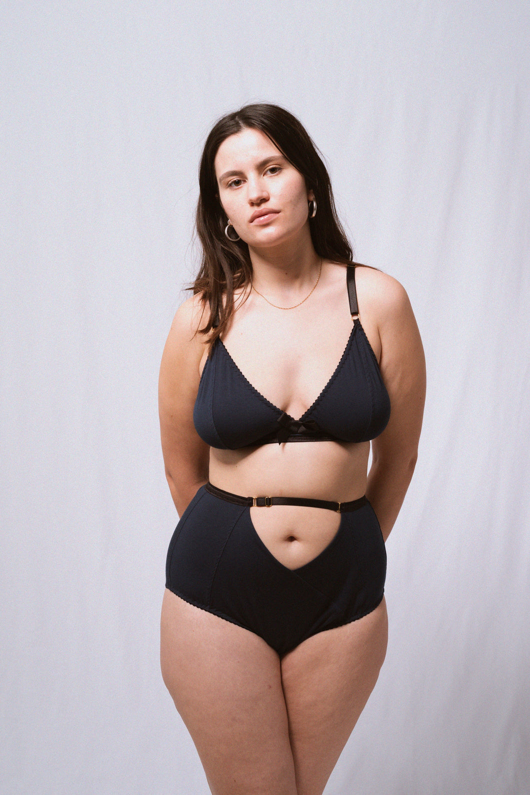 A long shot of a woman looking at the camera, wearing the Artemis bralette and high waist knickers. Both are in petrol blue bamboo jersey with black elastics and gold hardware. The front of the knickers has a cross over panel detail that creates an upside down triangle at the waistline around the belly button. The waistband carries on across the top of the triangle.