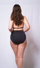 Cargar imagen en el visor de la galería, A full length image of a woman from the back. She wears the Artemis high neck bralette and high waist knickers. Her long brown hair covers the top of her back and the halter strap of the bralette. The back of the bralette is one adjustable strap across the back just below the shoulder blades. this has a gold slider in the centre. The knickers have a full back in petrol blue bamboo jersey.
