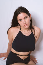 Cargar imagen en el visor de la galería, A woman looking up at the camera, she has long brown hair and gold hoop earrings. She wears the Artemis high neck halter bralette and high waist knickers in petrol blue bamboo jersey. The bralette comes up to just below her collar bones and has a scoop neck. The elastics on the bralette are black with gold hardware.
