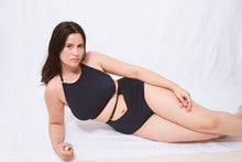 Cargar imagen en el visor de la galería, A woman lies on her side looking at the camera. She has long brown hair. She wears the Artemis high neck halter bralette and high waist knickers. The knickers have an upsidedown triangle cut out at the waistline around the belly button. The bralette sits just below her collar bones with a scooped neck.
