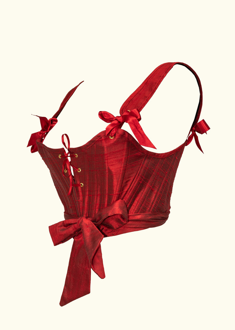 The side front of the red silk Olenska Stays. A lace up v at the centre front can allow the wearer to create a more curved bust shape. The eyelets are gold, and the straps attach with 2.5cm wide ribbon tied in bows.